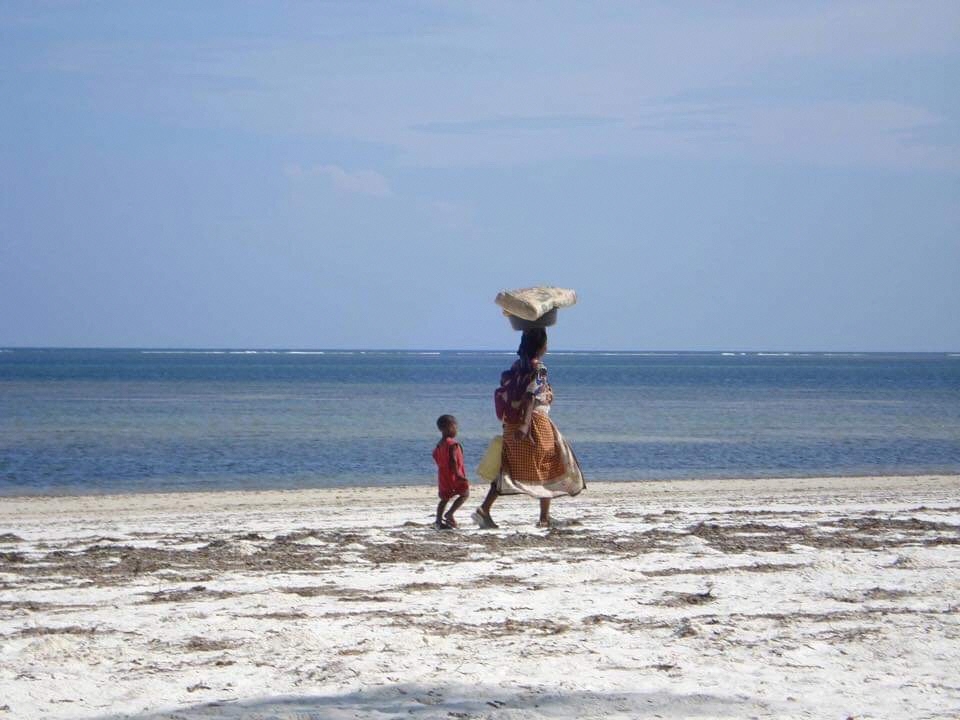 幼子をおんぶしながら頭に荷物を載せて歩く女性と一生懸命追いかける少年（ケニア共和国）